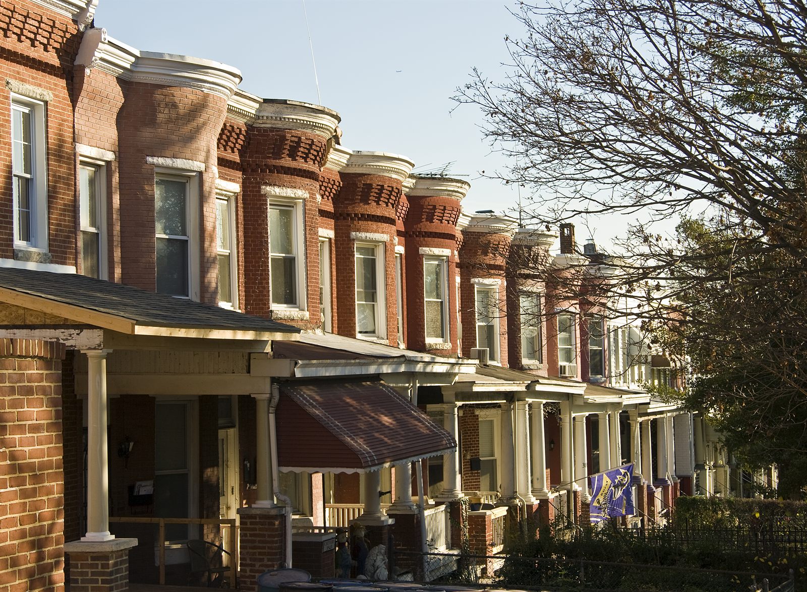 A row of two-storey red brick rowhouses receding down a Baltimore street in low afternoon sun. Each has a bay-fronted upper storey with decorative brickwork and a covered porch at ground level carried on white columns. Shallow front yards sit behind low fences, only a few feet deep, with the pavement immediately beyond them.